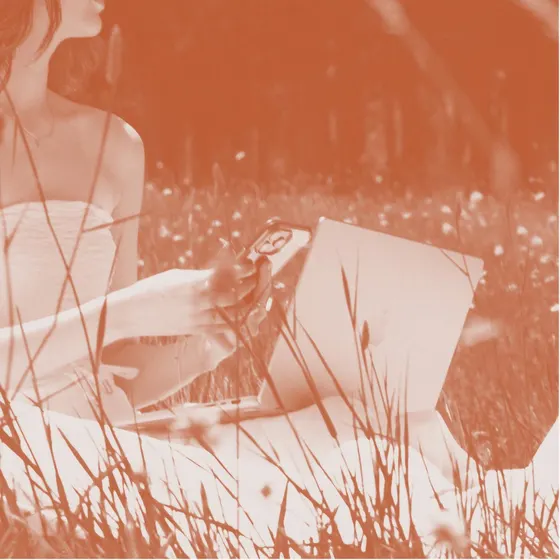 A woman holding a phone beside a laptop in long grass, toned in terracotta